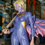 Cosplayer @thisoddangel dressed as a purple-skinned demon warrior with gold armor, lavender wings, and a sword, posing inside the Javits Center at New York Comic Con 2025.