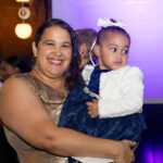 A woman in a gold dress holds her young daughter wearing a navy blue dress and white sweater at the CHiPS Annual Gala 2025 in Brooklyn.