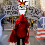 Protesters at the No Kings March in New York City, including one dressed in a penguin costume with an anti-ICE sign, another in a red cloak holding “Defend Democracy” and “We Will Not Be Silenced” signs, and a third holding a peace flag patterned like the U.S. flag, during the October 18, 2025 nationwide protest.