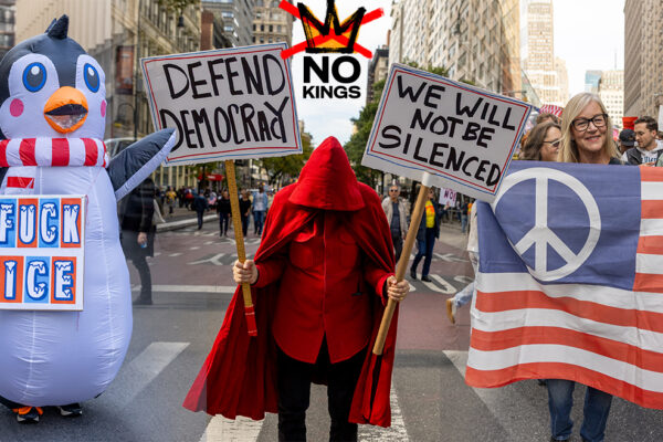 “Nearly 7 Million March in Historic ‘No Kings’ Rallies Across the U.S.” Protesters at the No Kings March in New York City, including one dressed in a penguin costume with an anti-ICE sign, another in a red cloak holding “Defend Democracy” and “We Will Not Be Silenced” signs, and a third holding a peace flag patterned like the U.S. flag, during the October 18, 2025 nationwide protest.