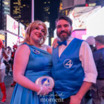 @harleynoxcosplay dressed as Sue Storm and Reed Richards, wearing blue formal Fantastic Four outfits with white “4” emblems, smiling and posing together at the New York Comic Con Time Square Meetup in Time Square, New York City.