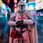 @themedievalmandalorian wearing white Mandalorian-style armor with a red cross emblem, chainmail, and cape, holding a sword and standing in Time Square at the New York Comic Con Time Square Meetup in New York City.