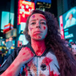 @jess__00 dressed as The Punisher, wearing a black tactical outfit with skull chest armor and realistic blood-effect makeup, standing and facing the camera at the New York Comic Con Time Square Meetup in Time Square, New York City.