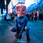 @mauricios_log dressed as Spider-Man “Bag-Man,” wearing a blue Fantastic Four bodysuit with the number 4 emblem and a paper bag mask, crouching at the New York Comic Con Time Square Meetup in Time Square, New York City.