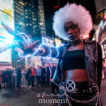 @mauricios_log dressed as Spider-Man “Bag-Man,” wearing a blue Fantastic Four bodysuit with the number 4 emblem and a paper bag mask, crouching at the New York Comic Con Time Square Meetup in Time Square, New York City.