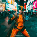 @sarahwood.cos dressed as Wolverine, wearing a yellow and black bodysuit with Wolverine mask, metal claws emitting a blue glow, holding a battle stance in Time Square at the New York Comic Con Time Square Meetup in New York City.