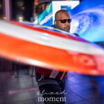 @gussyjay cosplaying as Captain America (Sam Wilson), wearing a red, white, and navy suit with sunglasses and swinging a large red-and-white Captain America shield in Times Square during the New York Comic Con Times Square Meetup.