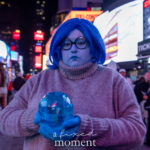 @howdoesonecosplay dressed as a blue Sadness-style character with blue face paint, blue wig, round glasses, oversized gray sweater, and blue gloves, holding a crystal-like orb while standing in Times Square during the New York Comic Con Times Square Meetup.