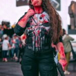@jess__00 dressed in a Punisher-inspired cosplay with a blood-splattered armored vest, black tactical pants, knee pads, and a handgun prop, standing in Times Square during the New York Comic Con Times Square Meetup.