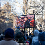 Crowd bundled in winter coats watching Zohran Mamdani speak on a large outdoor video screen during his January 1, 2026 inauguration outside New York City Hall.