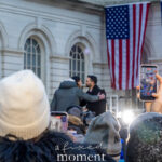 Zohran Mamdani embracing a supporter on stage outside New York City Hall during his January 1, 2026 inauguration, with American and New York City flags visible and a crowd recording the moment on smartphones.