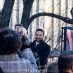 Zohran Mamdani speaking at a podium outside New York City Hall during his inauguration on January 1, 2026, with a crowd gathered and a large video screen displaying an American flag behind him.