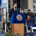 Zohran Mamdani speaking at a podium with the New York City seal outside City Hall during his January 1, 2026 inauguration, with officials seated behind him on stage.