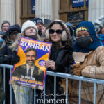 Supporters wearing winter clothing hold a Zohran Mamdani campaign poster behind metal barricades outside New York City Hall during the January 1, 2026 inauguration.