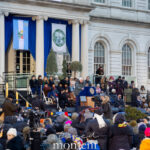 Wide shot of the Mamdani inauguration outside New York City Hall on January 1, 2026, showing the stage, seated officials, flags, media cameras, and a large crowd gathered in front.