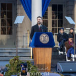 Zohran Mamdani speaking at a podium with the New York City seal outside City Hall during his January 1, 2026 inauguration, with officials seated behind him on stage.