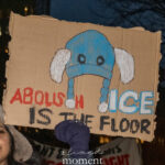Illustrated protest sign reading “Abolish ICE Is the Floor,” featuring a cartoon winter hat, held at a nighttime rally in New York City.