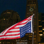 American flag waving at night with New York City buildings and a clock tower in the background.