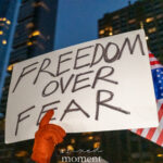 Handwritten protest sign reading “Freedom Over Fear” held aloft at night in New York City, with apartment buildings in the background.