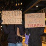 Two protesters holding signs that read “Kristi Noem is a Terrorist” and “Impeach + Indict Them All” at a nighttime demonstration in New York City.