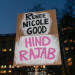 Hand-painted protest sign reading “Renee Nicole Good” and “Hind Rajab” in black and pink lettering, held at night in New York City.