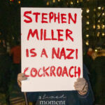 Hand-painted protest sign criticizing Stephen Miller, held at a nighttime rally in New York City.