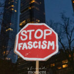 Red octagonal protest sign reading “Stop Fascism” held at night in New York City, with illuminated high-rise buildings in the background.
