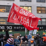 Red protest flag reading “No One Is Illegal” waved above a crowd during an anti-ICE march in New York City.
