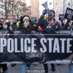 Protesters holding a large “Police State” banner while chanting through a crowded New York City street during an anti-ICE march.