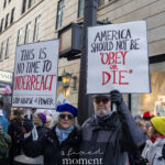 Two protesters holding signs that read “This Is No Time to Underreact” and “America Should Not Be ‘Obey or Die’” at the Hands Off protest on January 11, 2026, at 60th Street and Fifth Avenue in New York City.
