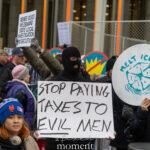 Masked protester holding a sign that reads “Stop Paying Taxes to Evil Men” at the Hands Off protest on January 11, 2026, near 60th Street and Fifth Avenue in New York City.