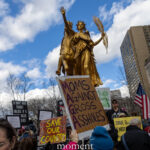 Protester holding a “Moms Against Gross Assholes” sign beneath a gold statue at the Hands Off protest on January 11, 2026, near 60th Street and Fifth Avenue in New York City.