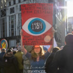 Protester holding a red “1984” sign with an eye illustration and a Trump quote at the Hands Off protest on January 11, 2026, in New York City.