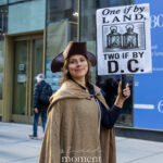 Protester wearing colonial costume holding “One if by Land, Two if by D.C.” sign at Hands Off protest in New York City.