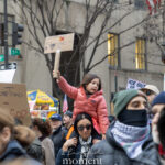 Child holding a handmade protest sign while sitting on an adult’s shoulders at the Hands Off protest in New York City.