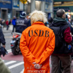 Back view of a protester wearing an orange prison jumpsuit marked “U.S.D.B.” at a New York City protest, with police and demonstrators in the background.