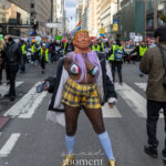 Protester wearing a Donald Trump mask and colorful protest outfit stands in the middle of a New York City street during a large political demonstration.