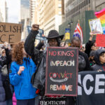 Protester raising a fist and holding a sign that reads No Kings Impeach Convict Remove during a large New York City demonstration.
