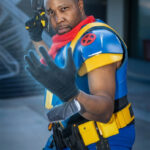 Bishop from X-Men cosplay wearing blue and yellow armored suit with red X insignia, red scarf, tactical belt, and holding a futuristic blaster at Dragon Con 2025 in Atlanta, GA. Cosplayer @coretoonzcosplay.