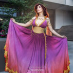 Megara cosplay in a magenta-to-peach ombré gown with gold trim and beige corset belt, holding the skirt wide at Dragon Con 2025 in Atlanta, GA. Cosplayer @blitzbun poses outdoors near greenery and steps.