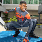 Miles Morales Spider-Man cosplay crouching on a blue metal table at Dragon Con 2025 in Atlanta, GA, wearing a black suit with red spider emblem, red shoulder accents, web-pattern detailing, and black sneakers. Cosplayer: @braxton.lee23.