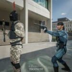 Solid Snake cosplay in a teal tactical bodysuit and black headband aims a prop pistol at a camouflaged guard during Dragon Con 2025 in Atlanta, GA. Cosplayer @nerd_alert_cosplay stages an action pose outdoors.