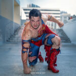 Battle-damaged Superman cosplay kneeling in superhero landing pose with glowing red eyes, torn blue suit, red cape, and distressed red boots at Dragon Con 2025 in Atlanta, GA. Cosplayer @supertotcosplay.