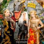 Three ACOTAR-inspired cosplayers—@lacyelizabethmcgee, @lucymason84, and @courtneyymichelleee—pose together in elaborate fantasy outfits in front of a large sculpted dragon head at the Monks Meadery display during Dragon Con 2025 in Atlanta, GA. One wears a gold gown and crown, one wears a black lace and velvet dragon-embroidered dress, and one wears a black leather High Fae outfit with white hair.