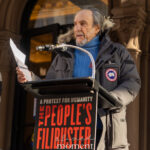 F. Murray Abraham reads from a sheet of paper while speaking at a lectern during The People’s Filibuster in New York City.