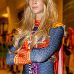 Cosplayer @sarahparrish.c dressed as Homelander from The Boys at Dragon Con 2025, wearing a blue patterned bodysuit, red gloves, gold belt, and a red-and-white cape. She stands with arms crossed, showcasing long blonde hair and confident expression.