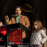 A speaker gestures at a podium during The People’s Filibuster outside The Public Theater, with a child standing nearby.