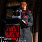 A speaker stands at a podium holding printed pages during The People’s Filibuster outside The Public Theater in New York City.