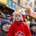 A child in a red traditional Chinese outfit and lion hat sits on an adultβs shoulders during the Lunar New Year Parade in Chinatown, New York City.