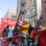 A parade performer in a yellow patterned jacket holds a confetti launcher while standing on a float during the Lunar New Year Parade in Chinatown, New York City.